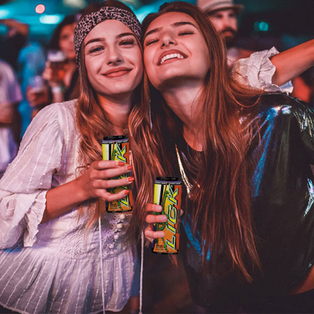 Two smiling women holding cans of LICK energy drink with a crowd in the background