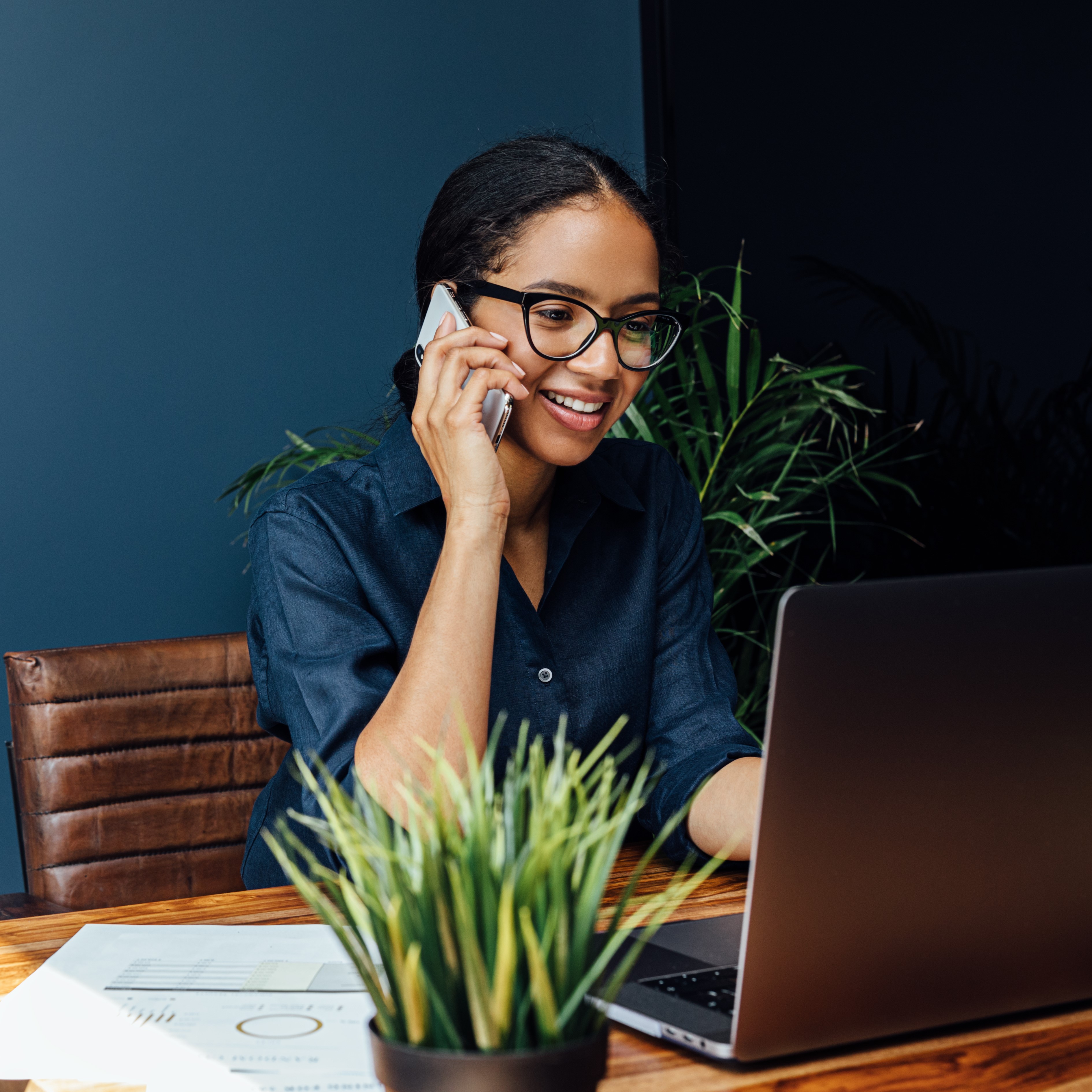 Woman sitting at a desk with a small plant, working on a laptop while talking on the phone