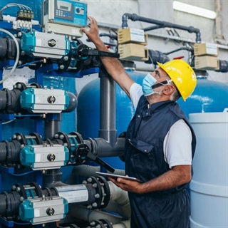Worker in a yellow hard hat, face mask, and vest looking at a system of tanks and display panel